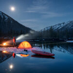 Person camping in a tent by a lake in the winter in the moutains at night.